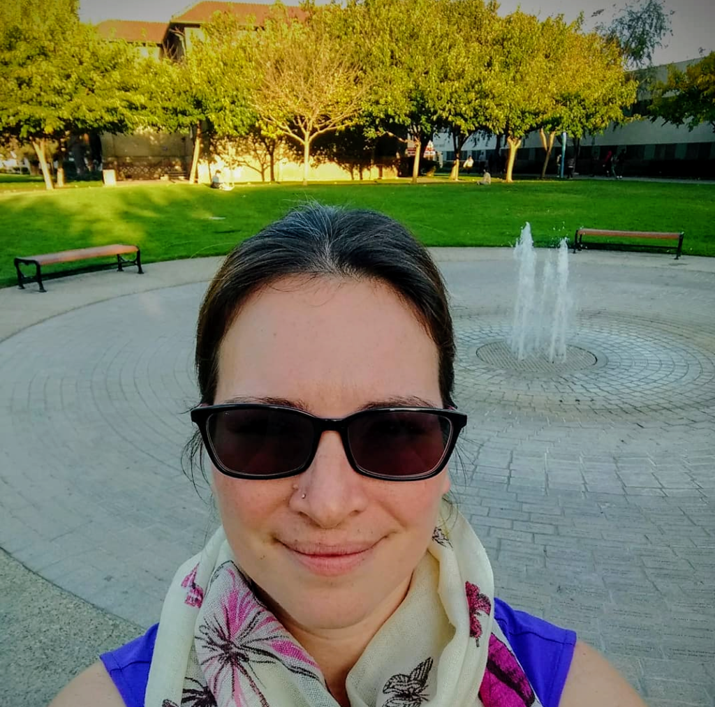 headshot of woman wearing sunglasses; flat water decorative water fountain, grass, and tress in the background