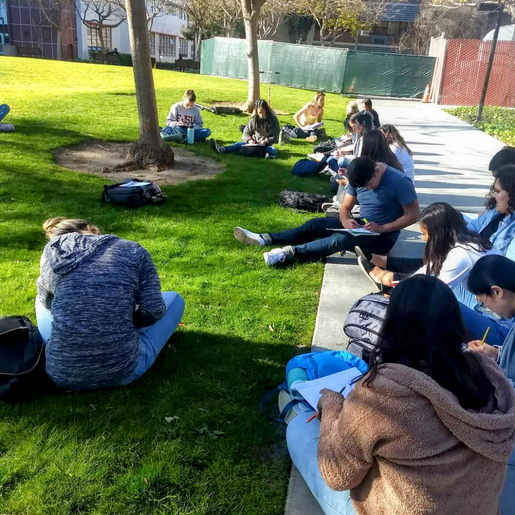 students experience the outdoors; a group of people sit on grass and walkway writing in notebooks